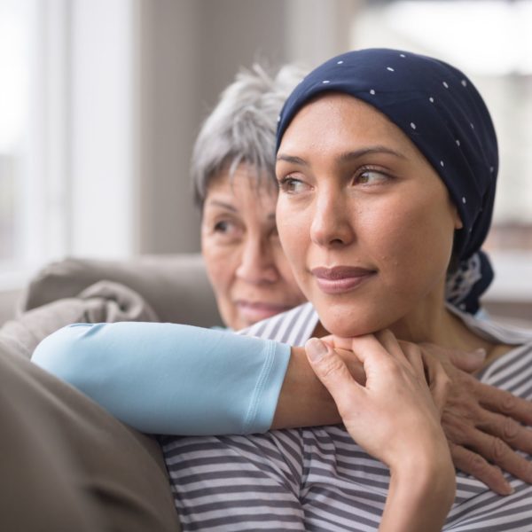 Mulher olhando para o lado com lenço na cabeça como uma pessoa que luta contra o câncers, endo abraçada por sua mãe.