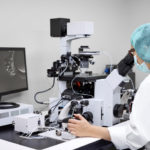 microscópio em laboratório de reprodução humana com uma pessoa realizando a biopsia embrionaria que aparece no monitor ao lado do microscópio