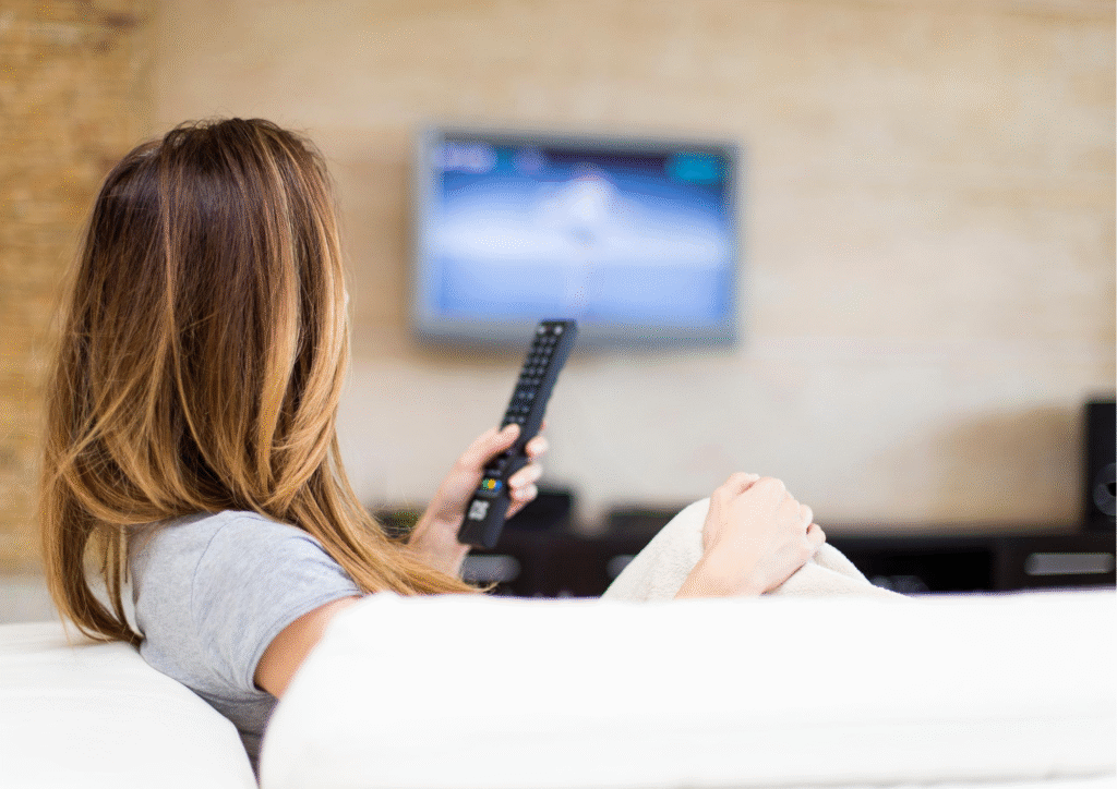 Mulher de costas sentada no sofá segurando o controle remoto da TV olhando para a tela onde passa um filme