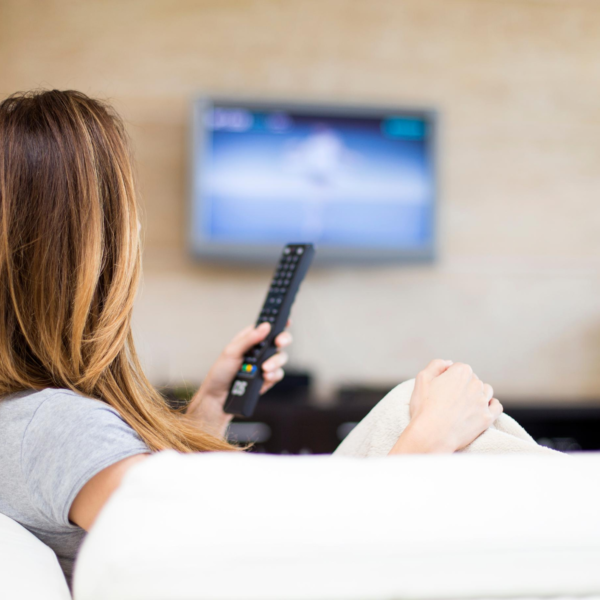 Mulher de costas sentada no sofá segurando o controle remoto da TV olhando para a tela onde passa um filme